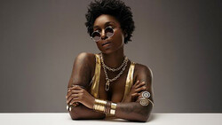 Portrait of a woman wearing round sunglasses, gold jewelry, and metallic body paint against a neutral gray background.