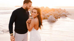 Couple embracing on a beach at sunset with rocky formations in the background.