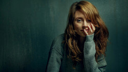 Woman with long brown hair resting her face against her hand, wearing a gray sweater against a teal background.