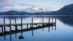 Wooden dock extending into still lake with snow-capped mountains reflected in water at dawn.