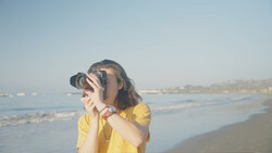 Woman in yellow shirt holding camera up to her face on a beach at golden hour.