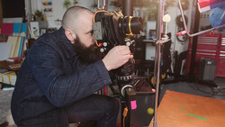 Filmmaker operating a vintage film camera mounted on a tripod in a workshop studio.