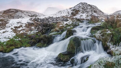 Cascading waterfall flowing over moss-covered rocks with snow-dusted mountains in the background.