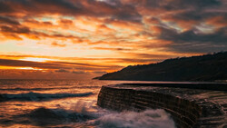 Dramatic coastal sunset with layered rock formations in foreground and mountainous headland silhouetted against golden sky.