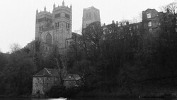 Gothic cathedral with twin spires rises above bare trees and a riverside building in monochromatic tones.