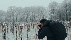 Photographer in black jacket shooting a frost-covered cornfield with bare trees in misty background.