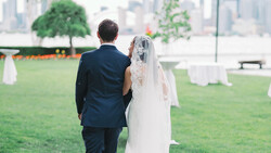 Bride and groom walking together on a grassy venue with a city skyline in the background.