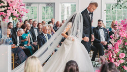 Groom helping bride with her veil during wedding ceremony indoors with pink floral arrangements.