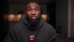 Man with beard wearing black hoodie looking directly at camera in indoor setting.