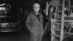 Older man in work clothes standing in a workshop or garage, black and white portrait.