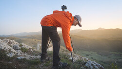 Hiker in orange jacket bending forward on rocky hilltop, adjusting camera mounted on head strap.