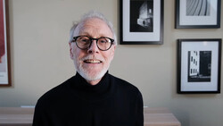 Older man with white hair and glasses wearing black turtleneck, seated indoors with framed black and white architectural photographs on wall behind.