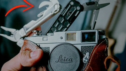 Photographer's hands holding a Leica rangefinder camera with brown leather strap, surrounded by vintage camera accessories and film photography gear.