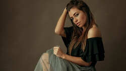 Woman in off-shoulder black top and gray skirt poses against neutral backdrop with hand to head.