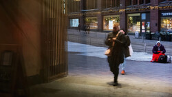 Woman in dark coat walking across an urban plaza at dusk, storefront windows visible in background.