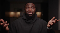 Man in black hoodie with hands raised, speaking directly to camera against dark background.
