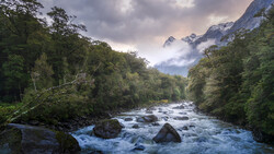 Rushing river with boulders flowing through forested valley beneath snow-capped mountains.