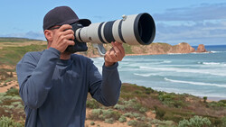 Photographer with telephoto lens positioned on coastal bluff overlooking rocky headland and ocean.