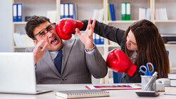 Two business professionals in an office playfully boxing with red gloves at a desk.