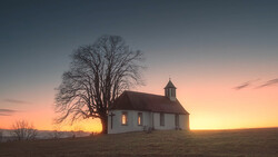 White wooden church with illuminated windows and bare tree silhouetted against golden sunset sky.