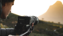 Photographer operating a mirrorless camera on a tripod during golden hour with mountains in the background.