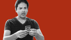Man in black v-neck t-shirt holding a smartphone, speaking expressively against a red background.