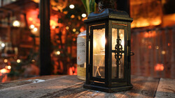 Black metal lantern with illuminated interior sitting on wooden surface with warm bokeh lights blurred in background.