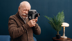 Older man with beard holding vintage film camera up to his face against blue background.