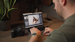 Photo editor working on a laptop displaying image editing software with a Canon camera and plant on wooden desk.