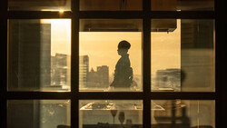 Silhouetted figure standing at a high-rise window overlooking a cityscape at golden hour.