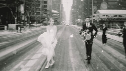 Bride in white dress running down urban street with groom and wedding party following behind.