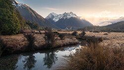 Snow-capped mountain peaks rise above a meandering stream in an expansive valley at golden hour.
