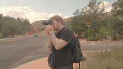 Photographer holding DSLR camera to eye while standing on roadside with mountains in background.