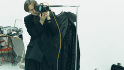 Photographer holding a camera to their eye while standing next to a clothing rack with a dark garment.