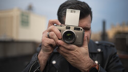 Photographer holding a rangefinder camera with flash attachment to his face while composing a shot.
