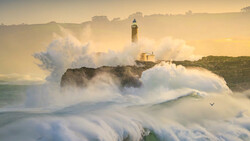 Lighthouse on rocky headland surrounded by massive crashing waves and sea spray at sunrise.