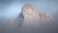 Snow-covered rocky peak emerging from thick fog and mist under overcast sky.