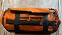 Orange and black cylindrical duffel bag with compression straps, displayed on a concrete surface.