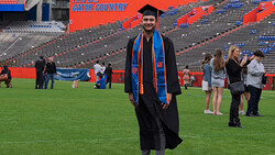 Graduate in cap and gown with blue and orange sash standing on stadium field during commencement ceremony.