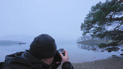Photographer shooting landscape at dawn by a misty lake with pine trees reflected in still water.