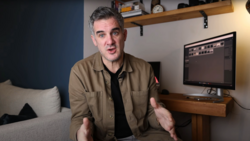 Man in tan jacket speaking to camera in home studio with monitor displaying editing software.