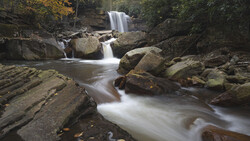 Cascading waterfall flowing over moss-covered rocks into a misty stream surrounded by dense forest.