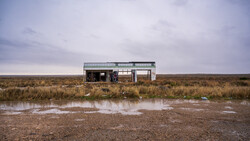 Abandoned single-story building with metal siding in vast, flat marshland under overcast sky.