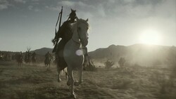 Silhouetted rider on horseback leading a group across an arid, mountainous desert landscape at sunset.