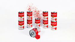 Campbell's tomato soup cans arranged in a group with one tipping over and dispersing red particles into the air.