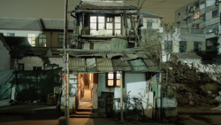 Weathered two-story residential building with illuminated doorway at night, surrounded by older structures.