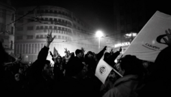 Nighttime crowd protest with raised hands and flags, urban buildings lit by street lamps.