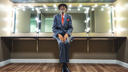 Person in blue pinstriped suit and red tie sitting on vanity counter beneath illuminated mirror with multiple bulbs.