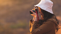Photographer with white bucket hat holding camera to face, backlit by warm golden light against blurred background.