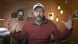 Man in burgundy quarter-zip pullover and white cap gestures expressively while speaking in a warm, wood-paneled interior space.